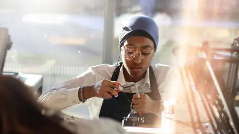 Aston University Engineering Academy A T-level jewellery student working at a bench making an item of jewellery. She has a headscarf around her neck and holds tools in her hands. She wears a white top with a black apron over it and is looking down at what she is working on.