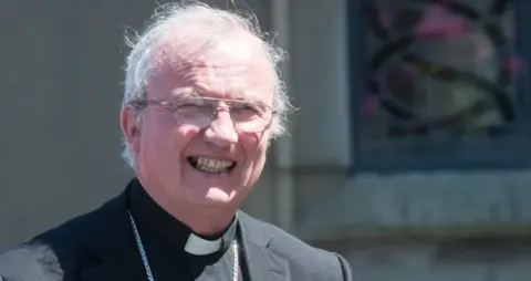 Pacemaker Donal McKeown is smiling at the camera. He is wearing a black jacket white and clerical collar with a large silver chain around his neck. He has white hair and gold glasses
