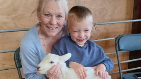 Family photo A smiling woman with light blonde hair, wearing a grey cardigan and sat on a black fold-out chair with a young smiling blonde boy wearing a blue jumper on her lap. On his lap is a white baby lamb, and both the woman and boy have their hands on the lamb.