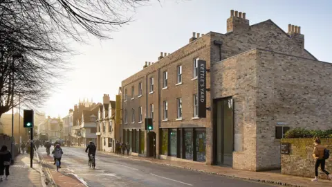 Jo Underhill View of Kettle's Yard from the street outside. The sun is setting and people are walking and cycling past the gallery. It is a brick building with black windows and a large black sign sticking out the front.