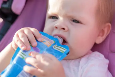 Getty Images A young toddler holds a blue-coloured food pouch to its lips and is eating from it while sitting in a pram