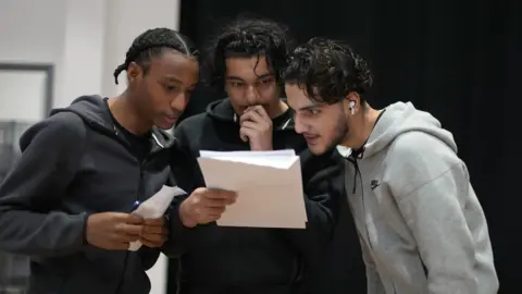 PA Three young men anxiously read the one in the middle's A-level results. He is holding his hand up to his mouth.