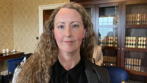 Children's Law Centre Rachel Hogan has light brown hair. She is wearing ear rings, a black shirt and a grey jacket. Behind her is a book shelf with wooden and glass doors, there are books inside on shelfs. There are blue chairs and a side table in the background. There is also a wooden door, with a white door frame behind her. 
