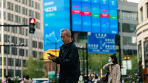 EPA A man looks at his phone in his hands as he walks down a street in Shanghai of high-rises with one building showing the results of the local bourse on its facade