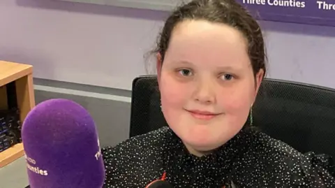 Justin Dealey/BBC Betsy Griffin is in the BBC 3CR radio studio sitting in a chair in front of a purple microphone. She is smiling at the camera and is wearing a sparkly black top.