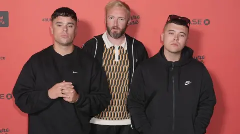 PA Media Mo Chara, left, is wearing a black sweatshirt and looking directly at the camera. He had short brown hair. DJ Provai, middle, is wearing a yellow and green patterned polo top and a black zipper jacket. He is looking directly at the camera and has a beard and ginger hair. Moglai Bap, is wearing a black zip up coat. He is wearing sunglasses on his head and has short brown hair.