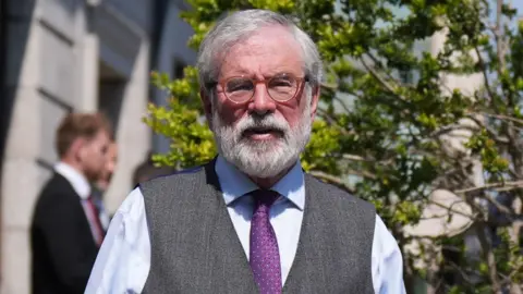 PA Media Gerry Adams, wearing a blue shirt, purple tie and grey waistcoat. He has grey hair and a grey beard and wears brown-rimmed glasses. The court is behind hin, with trees outside.