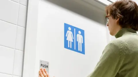 Getty Images Person walking into a toilet with a unisex gender sign on the door