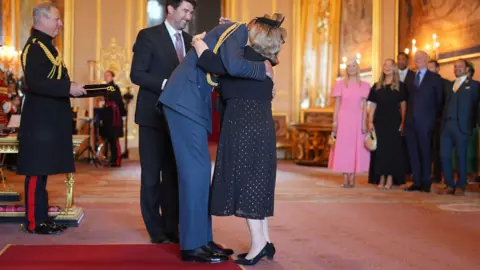 PA Media Wendy Daunt, wearing a navy mid-length dress with gold polka dots and a cropped tailored jacket with a fascinator hugs the Prince of Wales at an OBE investiture at Windsor Castle. Neither of their faces can be seen as they embrace as onlookers smile in the background.