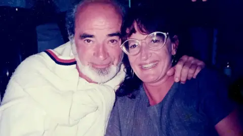 Family Eli Getreu is seen here with his wife Barbro Gentele. They are sitting close together and Mr Getreu has his arm around Ms Gentele. He is wearing a white top with a white jumper around his shoulders. She is wearing glasses and a blue dress and a watch.