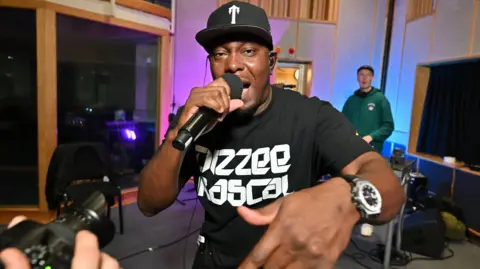 BBC Dizzee Rascal looks at the camera and is wearing a t-shirt with the words Dizzee Rascal on it. He is holding a microphone in one hand and wearing a black baseball cap.