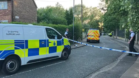 BBC Merseyside Police officers and vehicles inside a cordon following an incident around the Tobruck Road area in Huyton on a sunny evening.