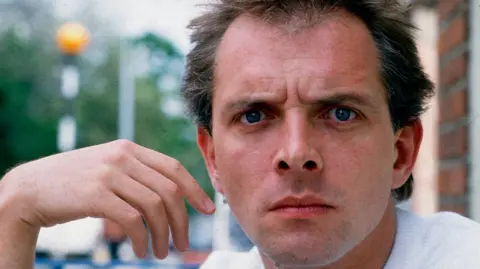 Steve Rapport/Getty Images Rik Mayall, a clean-shaven man with dark hair, looks at the camera, glaring at the camera in a light-hearted way. His hand is raised and hangs limp by the side of his face.