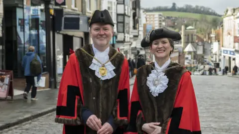 BBC A man and a woman on a cobbled high street wearing red and brown robes and historical white collars. They are also wearing traditional black hats and mayoral medallions.