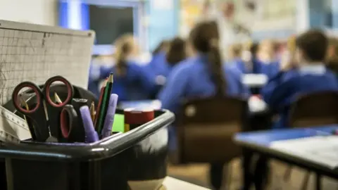PA Media A box of stationery - including a whiteboard, pens and scissors - with a class of primary school students in the background.