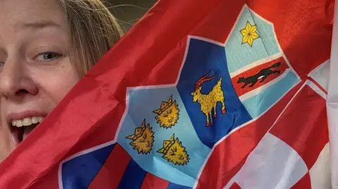 Emma Gale Emma Gale with her mouth wide in an excited expression, holding up a Croatian flag which is partly obscuring her face