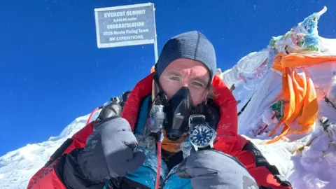 Blue Door Productions/Stan Gaskell Man in breathing gear and very warm, padded clothing for mountain expedition gives the thumbs up- he is wearing very padded gloves, the sun is shining and the mountain and bits of material and a flag are seen behind him.