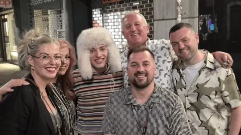 Scott Ross/OGAE UK Members of the British Eurovision fan club OGAE UK pose for a photo with Eurovision 2024 winner Nemo. Nemo is wearing a cream, fluffy hat and stripped shirt. Everyone is smiling for the photograph.