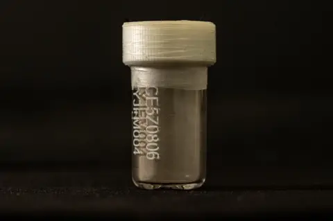 Tony Jolliffe/BBC News A small clear vial with a white plastic top and tape around the lid. It is lit against a black background. On the front is a code. Inside at the bottom there is a coating of dust.