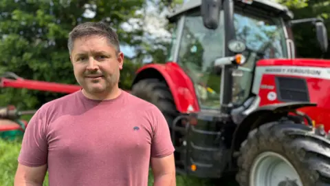 BBC A man in a pink t shirt stands in front of a red tractor. They are in a field getting ready to cut grass.