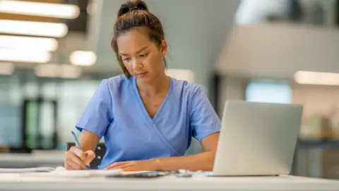 Getty Images Nurse in a blue uniform, writing at a desk, with a laptop computer in front of her.