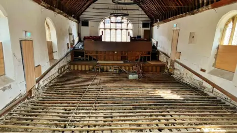 PA Media The interior of a small white painted theatre with exposed wooden floor supports and base leading towards a wooden stage. The ceiling is arched and has wooden beams, and windows going around the building. 