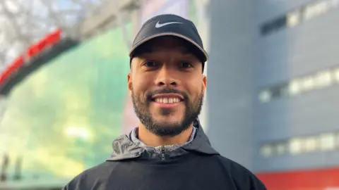 BBC Joe Thompson, wearing a baseball cap, smiles in front of Manchester United's stadium