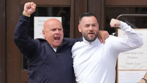 PA Media Two men with their arms around each others shoulders with their hands raised smile outside a court building