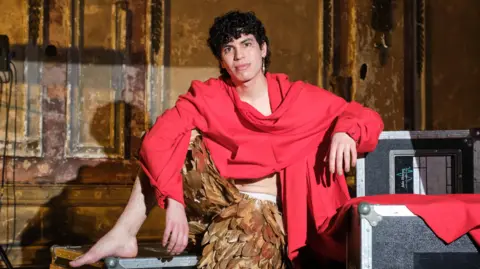 BBC Jacob Alon, wearing gold feathered trousers and an oversized red sweater, poses on flight cases backstage at the BBC's Jools Holland show