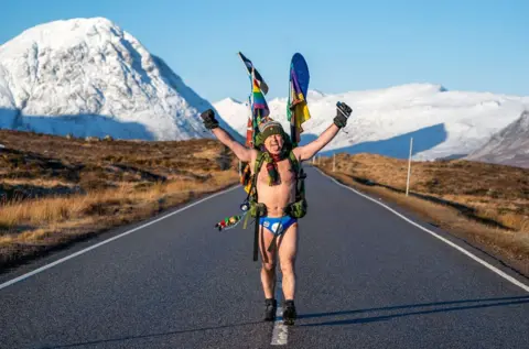 PA Media Mick Cullen, otherwise known as Speedo Mick, walks through Glencoe in sub-zero temperatures wearing only his swimming trunks. He is wearing his blue Speedos, a woolly hat, scarf, backpack, gloves and nothing else, his face tilted upwards to the sky with his eyes closed and a look of relief, against a background of snowy mountains in Glencoe, Scotland, in January 2023