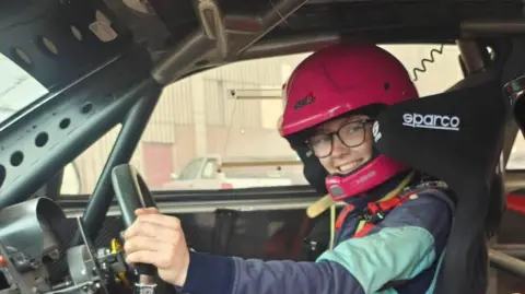BBC Letisha Conn is sitting in the drivers seat of a rally car, she is holding the gear stick with one hand. She is wearing a pink helmet and glasses. Another car can be seen out of the drivers window. She is also wearing navy and green over all's. She is sitting in a black seat with white writing. The car also has a roll cage within its interior.