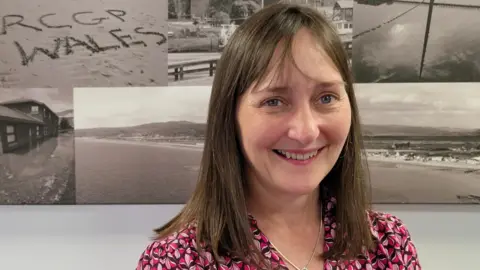 RCGP Dr Rowena Christmas has brown shoulder length hair and a partial fringe. She has blue eyes and is smiling at the camera. She is wearing a patterned shirt with different shades of pink and is stood in front of some black and white beach photos