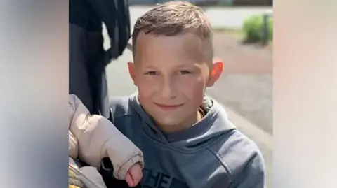 Family photo Layton Carr is smiling into the camera. He has short brown hair, cropped at the sides, and is wearing a grey hooded top. The arm of a baby in a puffer jacket is in front of him to his left.