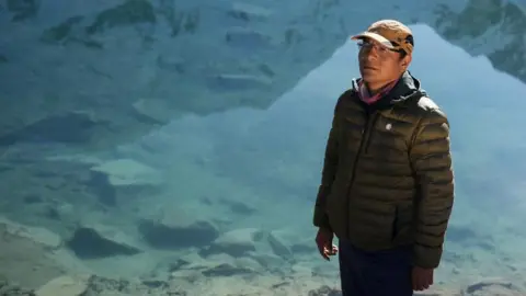 Reuters Peruvian farmer Saul Luciano Lliuya poses for a photo in front of Lake Palcacocha. He is wearing a baseball cap and behind him the clear waters of the lake can be seen. 