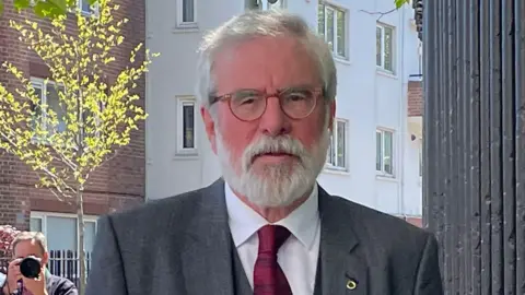 PA Gerry Adams is pictured from the chest up - he wears a grey jacket, grey waistcoat and red tie. He has two pins on his lapel. He has grey hair, a grey beard and brown glasses. In the background you can see a photographer taking a picture.