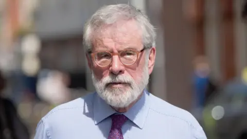 PA Media Gerry Adams walks outside the High Court in Dublin. He has short white hair and beard. He is wearing small, orange circular glasses and his expression is neutral. He is also wearing a light blue collared shirt and a purple patterned tie.