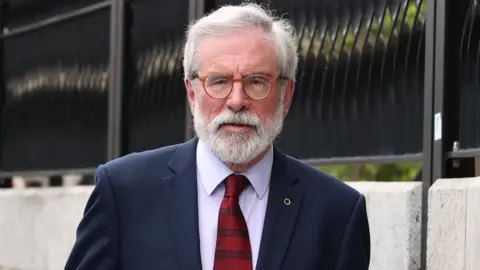 PA Media Gerry Adams has white hair and a beard. He wears brown glasses, a navy suit, a light shirt, a red tie, and a pin on his lapel