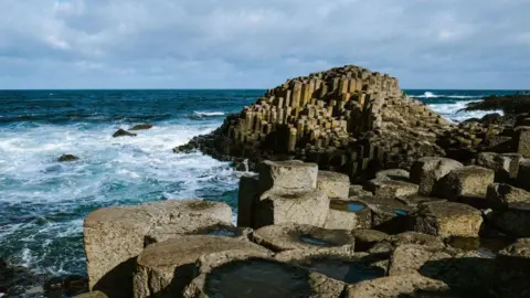 Getty Images Hexagonal columns all fitted together at the edge of the coast. The sea is rough and has white foam at the rocks. 
