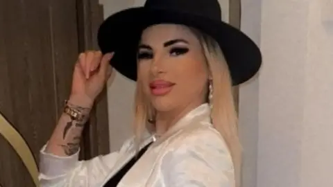 Garda A woman, with blonde hair, wearing a black hat and white shirt. She is holding the hair with one hand. She has tattoos on this arm. She is smiling at the camera.