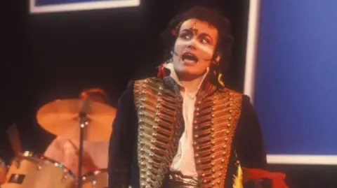 BBC Adam Ant on stage in the 1980s with a drum kit behind him. He is wearing a gold military-style jacket with lots of buttons and brocade and has his traditional white stripe painted across his nose and upper cheeks. He appears to be mid song.