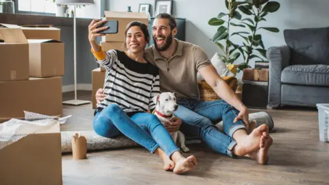 Getty Images Couple sitting on a rolled up carpet with their dog take a selfie in their new home surrounded by cardboard boxes.