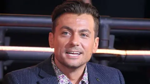 Getty Images Paul Danan wearing a blue pinstripe blazer over a pink flowery shirt. He has blue eyes and dark hair which is swept backwards and shaved short on the sides.