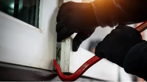 Getty Images Two gloved hands using a red crowbar to break open a window, which is painted white.