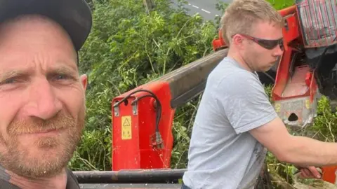 CPS Daniel Graham, wearing a black baseball cap and with a ginger beard, takes a selfie of him and Adam Carruthers, who is wearing sunglasses and a grey shirt, with short fair hair, and is cutting at a tree with a chainsaw