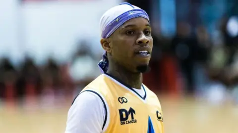 Getty Images A man wears a yellow basketball jersey over a white t-shirt, and a blue-and-white bandana covering his head. A section of basketball court and a row of spectators can just be made out in the background.