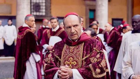Alamy A group of clergy members dressed in ornate liturgical vestments, including red and gold embroidered chasubles and matching mitres, are gathered outside a building with columns. In the middle is Ralph Fiennes as Cardinal Lawrence, dressed in full liturgical vestments.