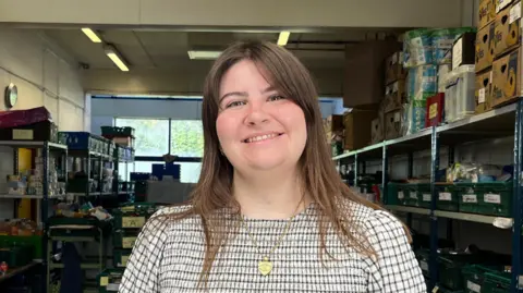 Cambridge City Foodbank Kate McIntosh is smiling at the camera while in the food bank. She is wearing a blue and white checkered top and a gold necklace. She has long brown hair and a side fringe. Behind her are shelves with green trays on. In the centre of the room, also behind her, are more trays stacked up with food inside them.