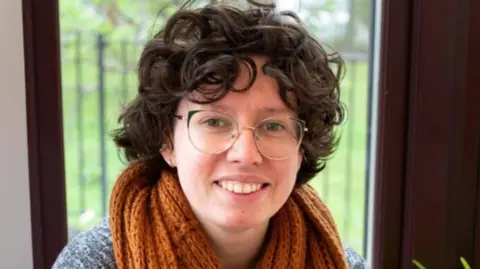 Jenny Watt Jenny Watt smiles while looking directly into the camera. She has glasses, curly dark hair and an orange/brown scarf wrapped around her neck.
