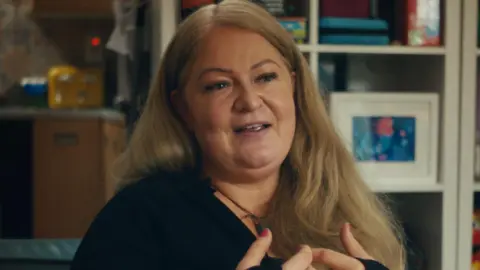 BBC/Alleycats TV Lynn McDonald with long, blonde hair sits on a sofa. She wears a black top. There is a bookcase behind her with books and photoframes.