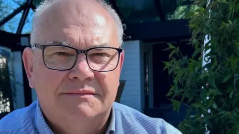 BBC A man in a blue shirt and glasses looks at the camera